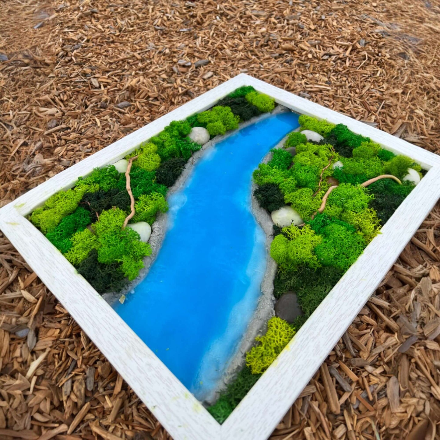 Epoxy and moss wall art with a sky-blue epoxy river flowing through textured moss and natural rocks.  The artwork rests on a bed of wood chips, highlighting the natural elements and handcrafted details